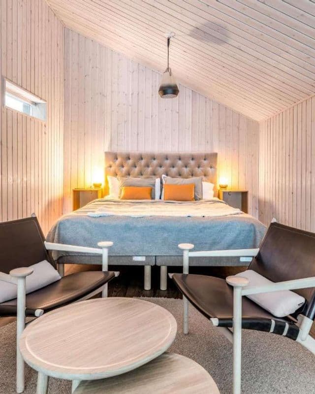 Una camera da letto moderna con pareti rivestite in legno chiaro e un soffitto a volta, con un letto capitonné e due poltrone in pelle nera.