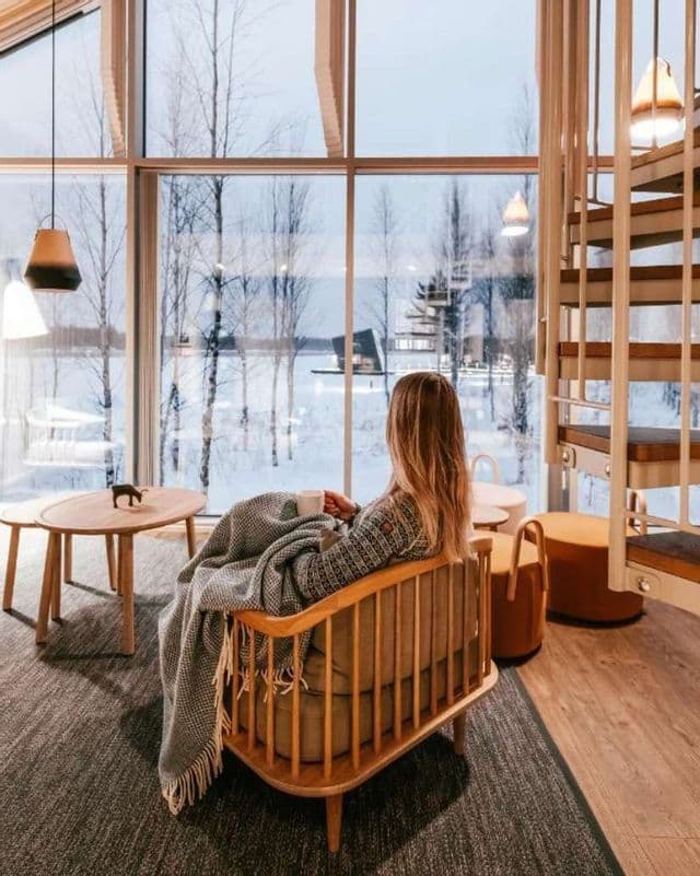 Una donna seduta in una poltrona di legno, avvolta in una coperta, sorseggia da una tazza mentre ammira un paesaggio innevato da un'ampia finestra.