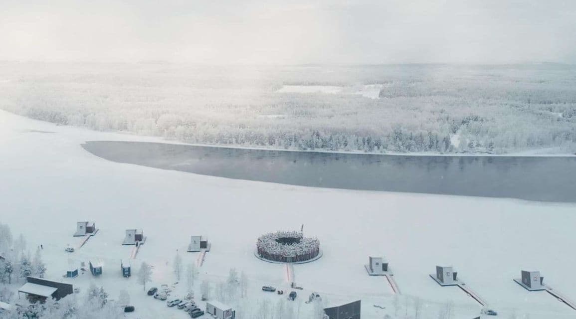 Veduta aerea di un resort con baite e un edificio spa circolare sulla riva di un lago parzialmente ghiacciato in un paesaggio innevato.