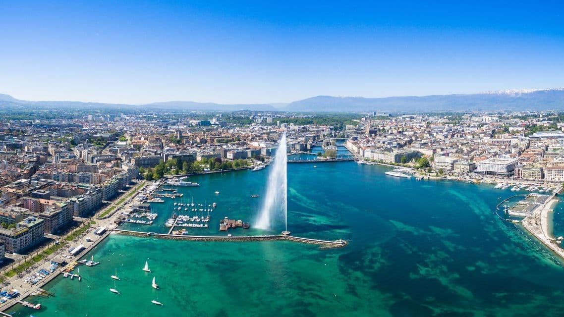 Vue aérienne d'une grande fontaine jaillissant de l'eau d'une baie turquoise, avec un paysage urbain dense et des montagnes lointaines.