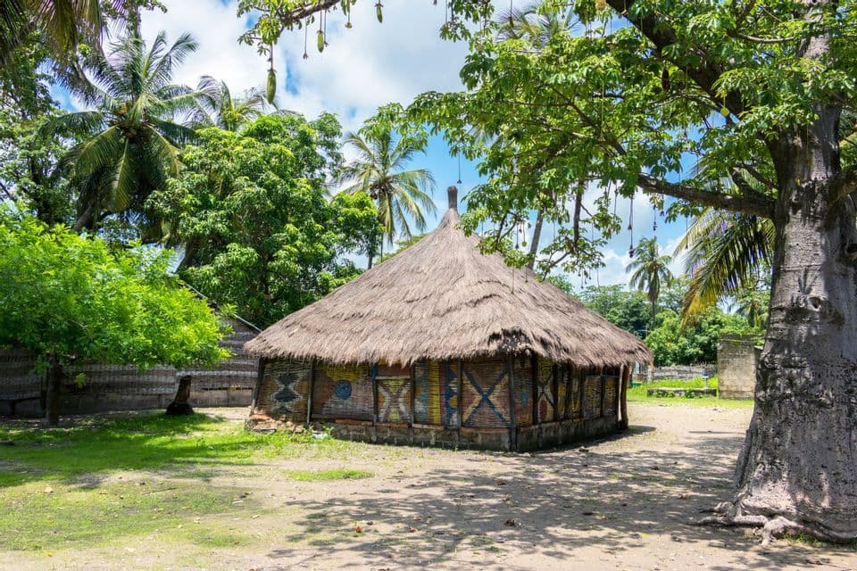 Una cabaña tradicional con techo de paja y paredes pintadas de colores se alza en un claro entre exuberantes árboles tropicales y palmeras.