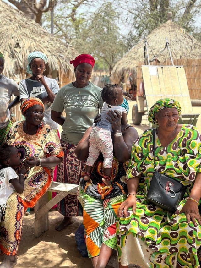 Un grupo de mujeres y niños con ropa colorida y estampada se reúne al aire libre en un pueblo con chozas de techo de paja.