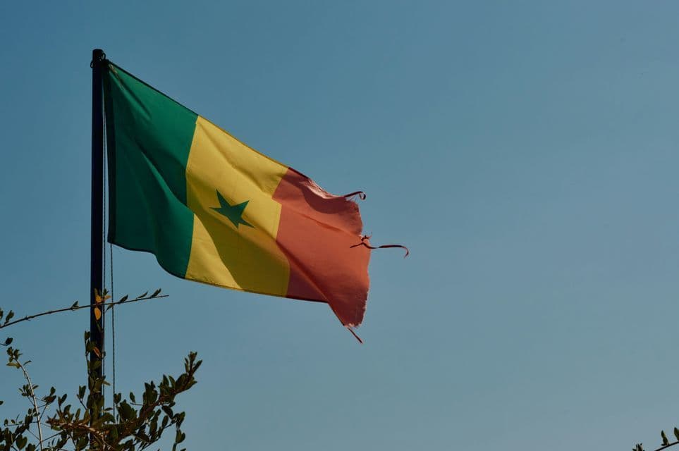 La bandera de Senegal, con un borde deshilachado, ondea en un mástil contra un cielo azul claro.