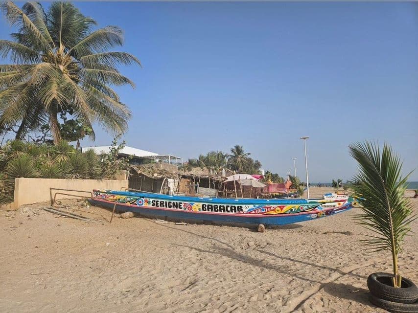 Un barco de madera largo y de colores brillantes con el nombre "Serigne Babacar" descansa en una playa de arena junto a palmeras bajo un cielo despejado.