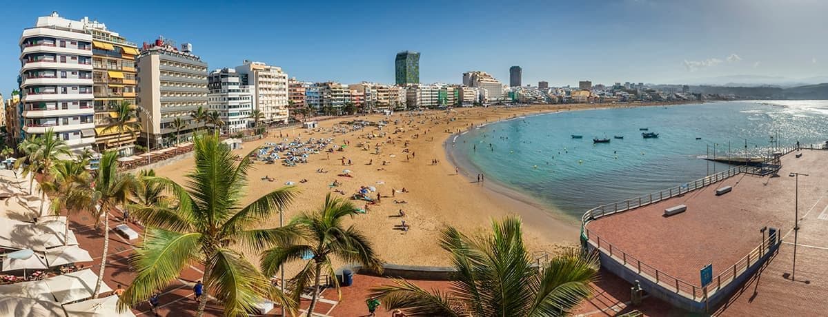 Vista panoramica sopraelevata di una spiaggia cittadina affollata, con persone che prendono il sole e nuotano, fiancheggiata da edifici e palme.