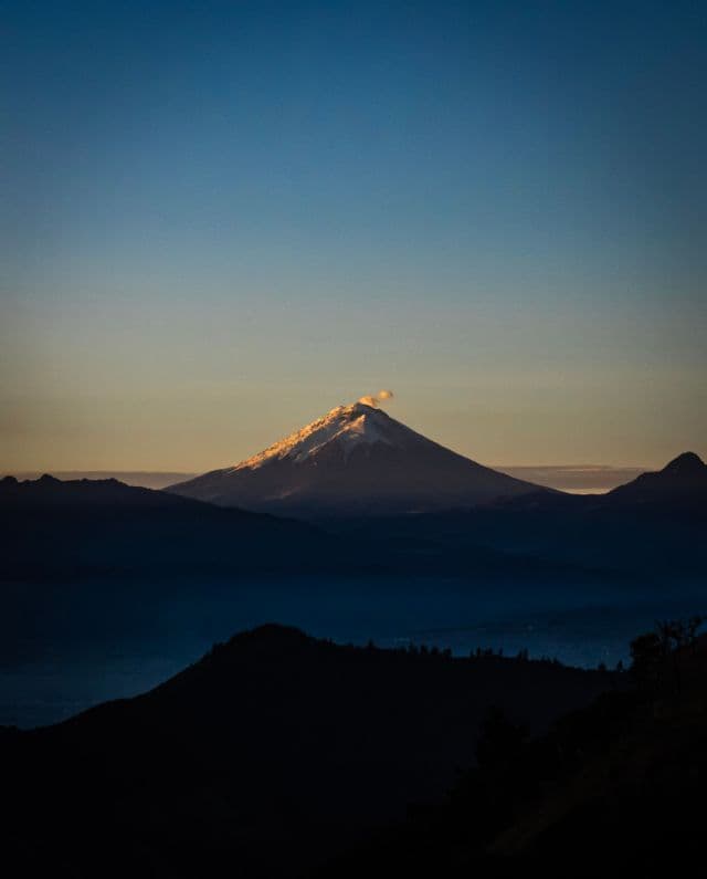 A snow-capped volcano's peak, lit by golden sunrise light, emits a plume of smoke above layers of dark, silhouetted mountains.