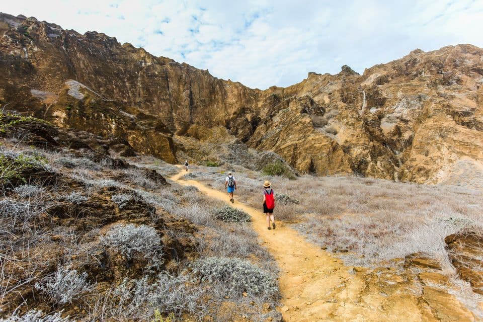 A WeRoad group trip hikes along a narrow dirt path through a rocky valley, with steep cliffs towering in the background.