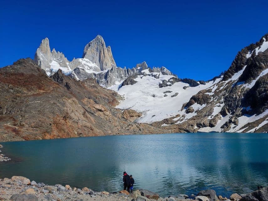 Due persone di un viaggio di gruppo WeRoad si trovano su una riva rocciosa, guardando un lago turchese e montagne innevate.