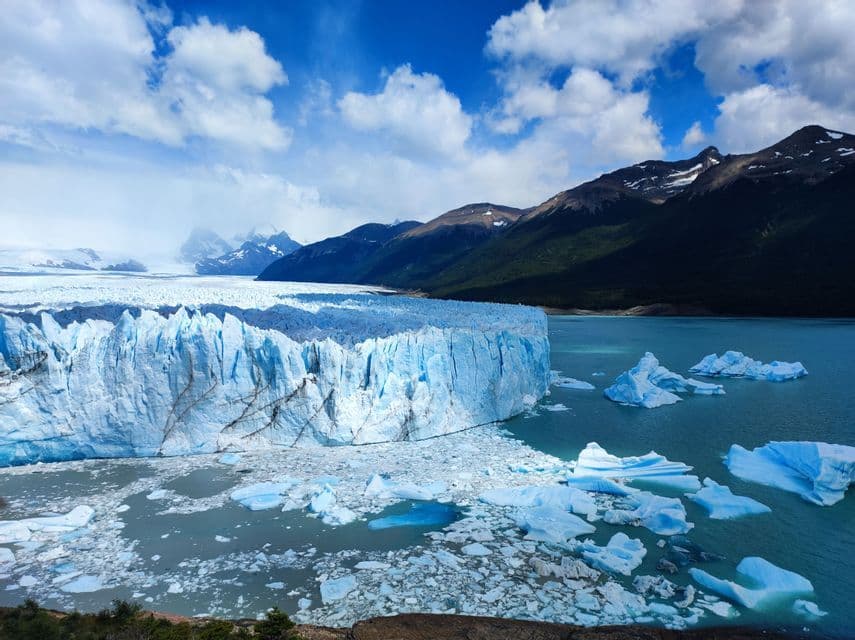 Un enorme ghiacciaio rilascia iceberg in un lago di fronte a una catena montuosa sotto un cielo parzialmente nuvoloso.