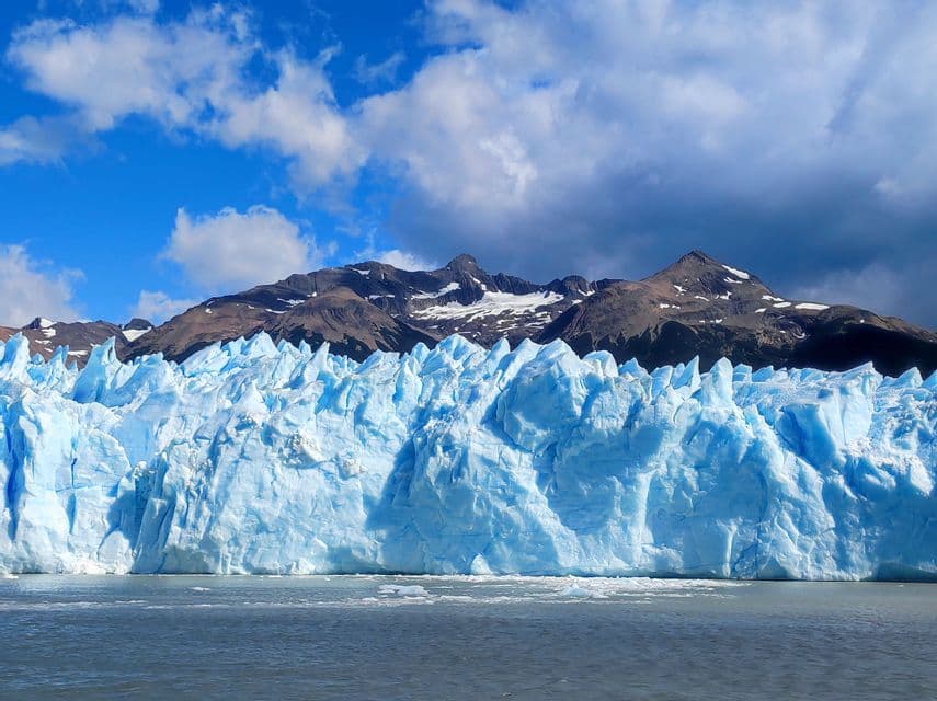 Un grande ghiacciaio azzurro si erge in acque tranquille di fronte a un'aspra catena montuosa sotto un cielo parzialmente nuvoloso.