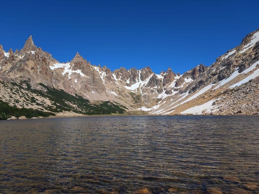Una vista dal basso di un limpido lago alpino con una catena montuosa frastagliata e rocciosa innevata sotto un cielo azzurro intenso.