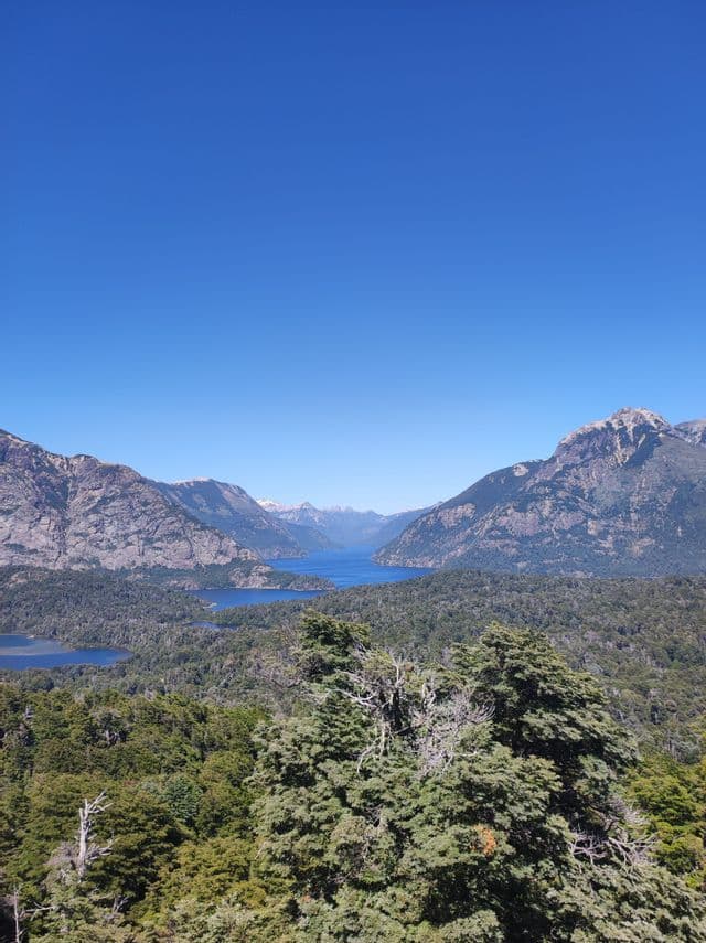 Una vista panoramica di un grande lago blu circondato da montagne boscose sotto un cielo sereno.