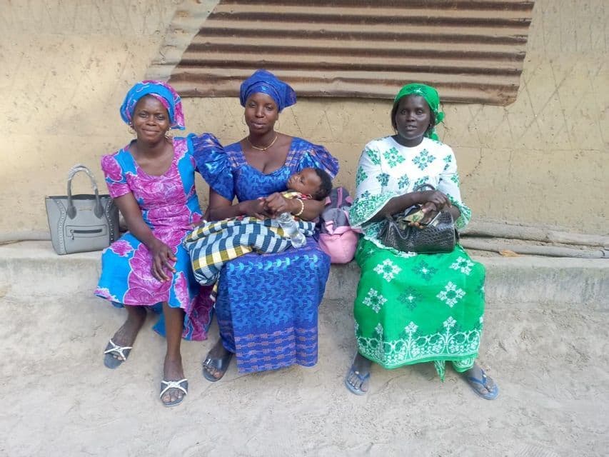 Tres mujeres con coloridos atuendos tradicionales se sientan en un escalón al aire libre, una de ellas con un bebé dormido en su regazo.