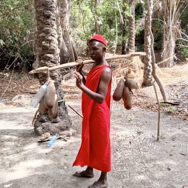 Un hombre con túnica y gorro rojos está de pie en una zona boscosa, llevando un palo sobre sus hombros con calabazas colgando de él.