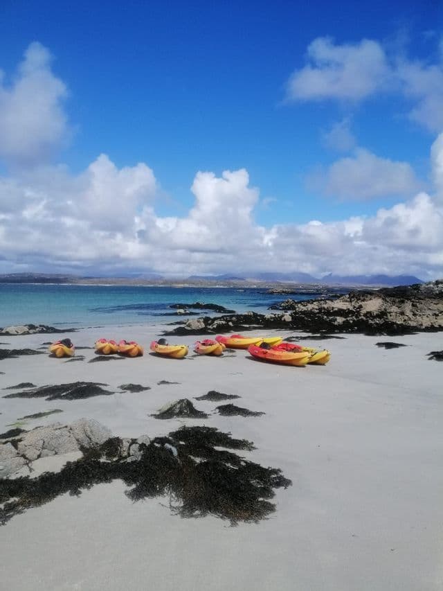 Plusieurs kayaks colorés sont garés sur une plage de sable blanc avec des algues sombres, surplombant une baie turquoise sous un ciel bleu nuageux.