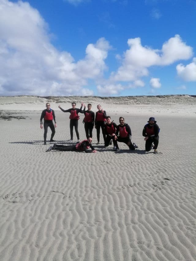 Un voyage de groupe WeRoad, en combinaisons de plongée et gilets de sauvetage rouges, posant pour une photo sur une vaste plage de sable.
