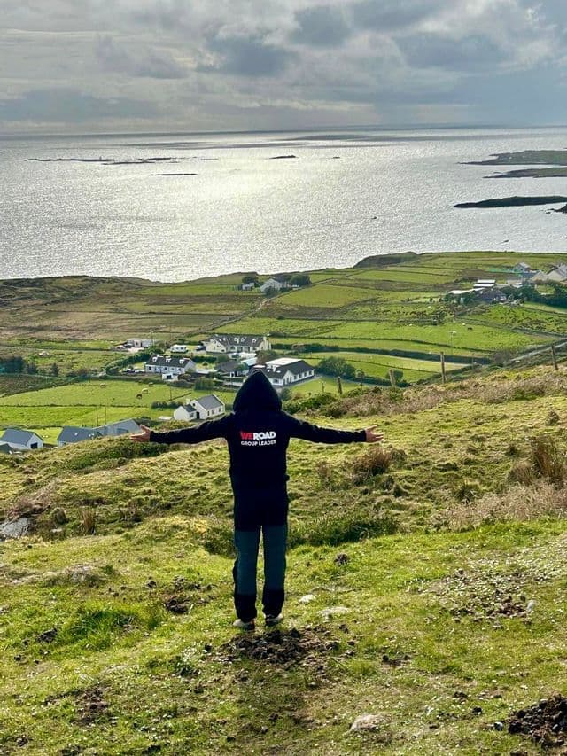 Un chef de groupe WeRoad les bras tendus sur une colline herbeuse, surplombant un village côtier et la mer.