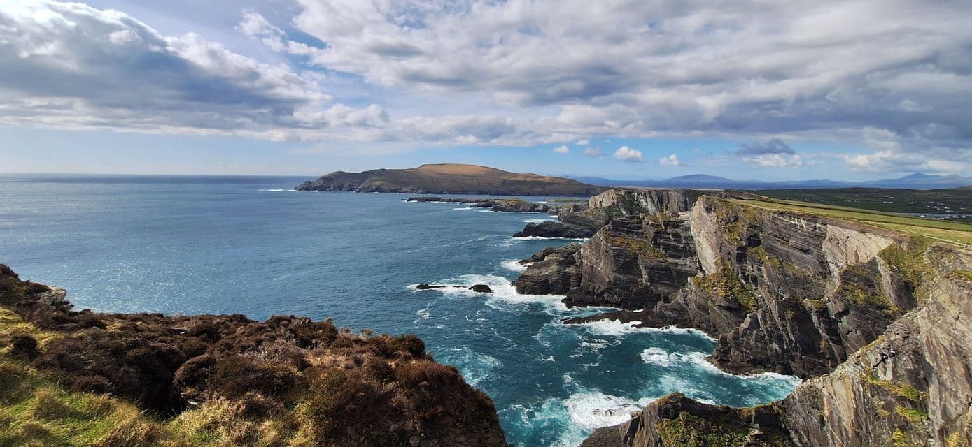 Une côte déchiquetée avec des falaises rocheuses abruptes et stratifiées plongeant dans un océan bleu agité sous un ciel partiellement nuageux.
