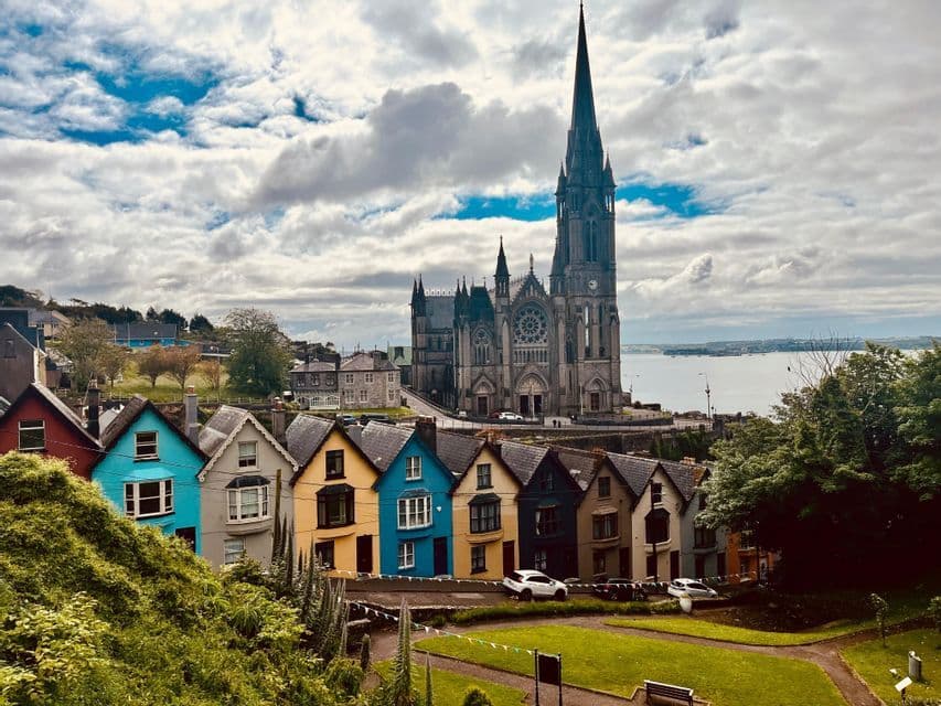Une rangée de maisons colorées se trouvent sur un flanc de colline, sous une grande cathédrale, surplombant un plan d'eau sous un ciel nuageux.