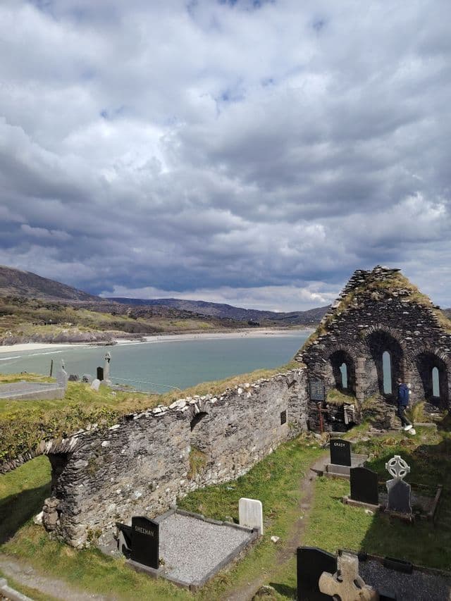 Ruderi in pietra di una chiesa e un cimitero che si affacciano su una baia con spiaggia sabbiosa e colline sotto un cielo nuvoloso.