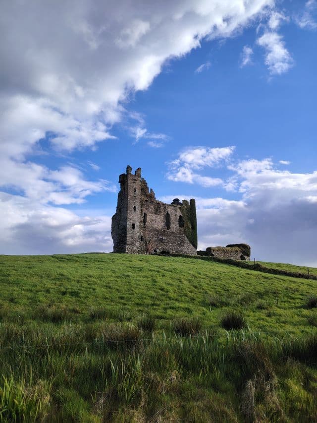 Le rovine di un castello di pietra si ergono su una rigogliosa collina verde sotto un cielo azzurro con nuvole bianche.