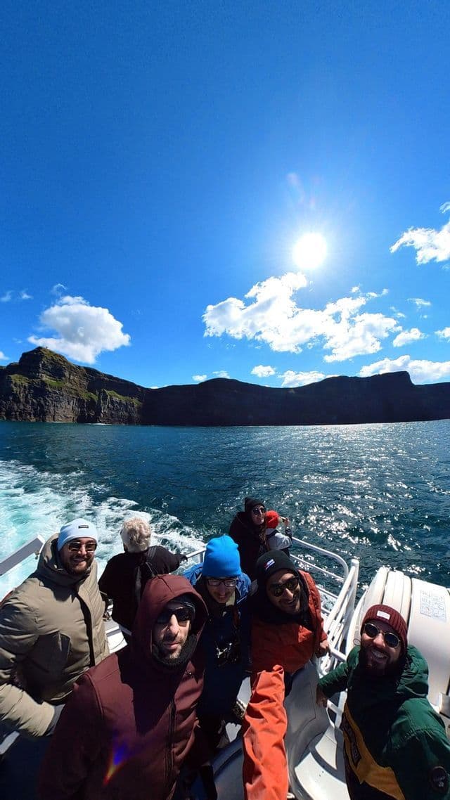Un voyage de groupe WeRoad prend un selfie sur un bateau naviguant le long de falaises rocheuses sous un soleil éclatant et un ciel bleu.