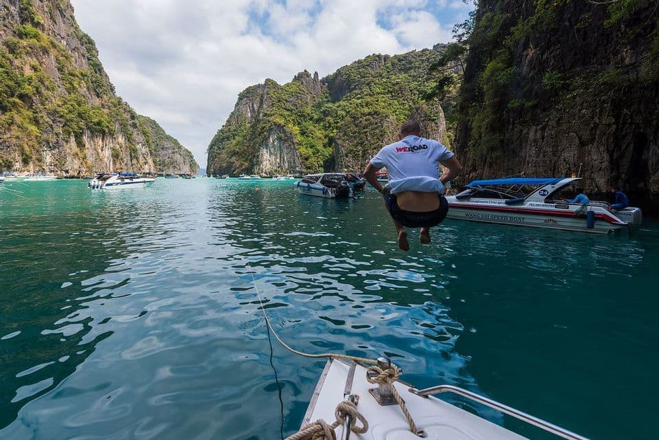 Un hombre con una camiseta de WeRoad salta de un barco a las aguas turquesas de una laguna rodeada de acantilados rocosos.