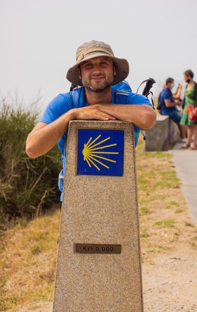 Ein lächelnder Mann mit Hut und Rucksack lehnt an einem Steinmarker, der 'Km 0,000' mit einem gelben Muschelsymbol anzeigt.