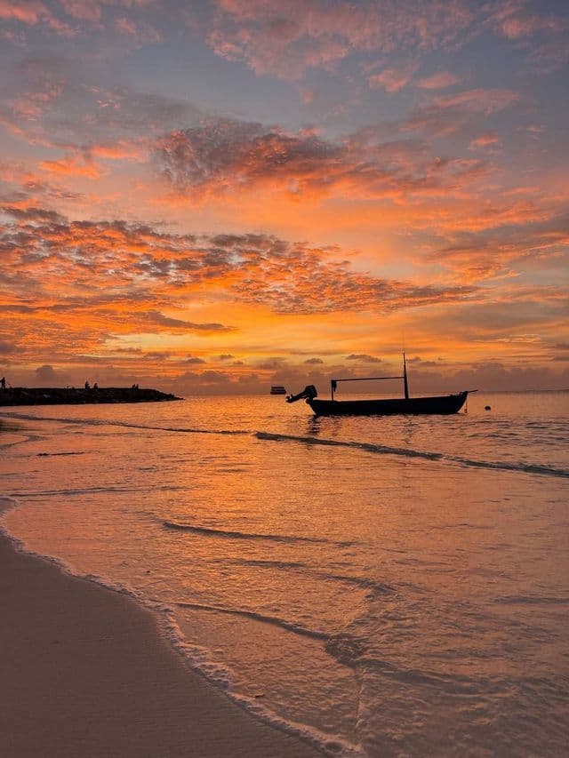 Ein silhouettiertes Boot schwimmt auf dem Meer nahe einem Sandstrand unter einem leuchtend orangefarbenen Himmel bei Sonnenuntergang.