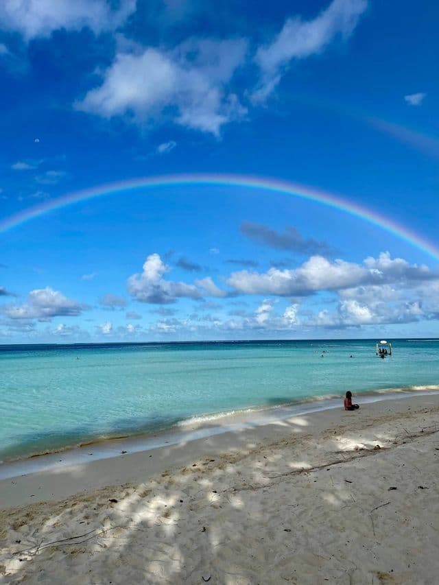 Un grande arcobaleno si inarca su una spiaggia tropicale con acqua calma e turchese mentre una persona è seduta sulla sabbia.