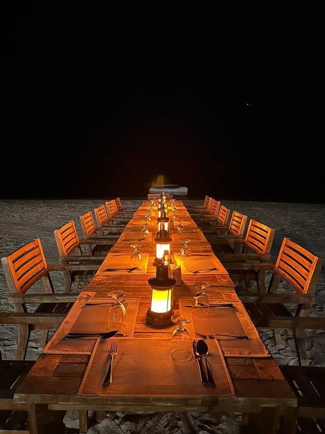 Una larga mesa de madera con servicios y linternas está preparada para la cena en una playa de arena por la noche.