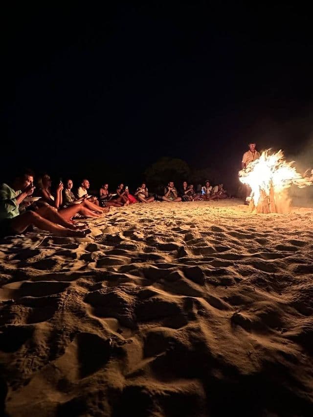 Eine WeRoad-Gruppe sitzt nachts an einem Sandstrand, versammelt im Kreis um ein großes Lagerfeuer.