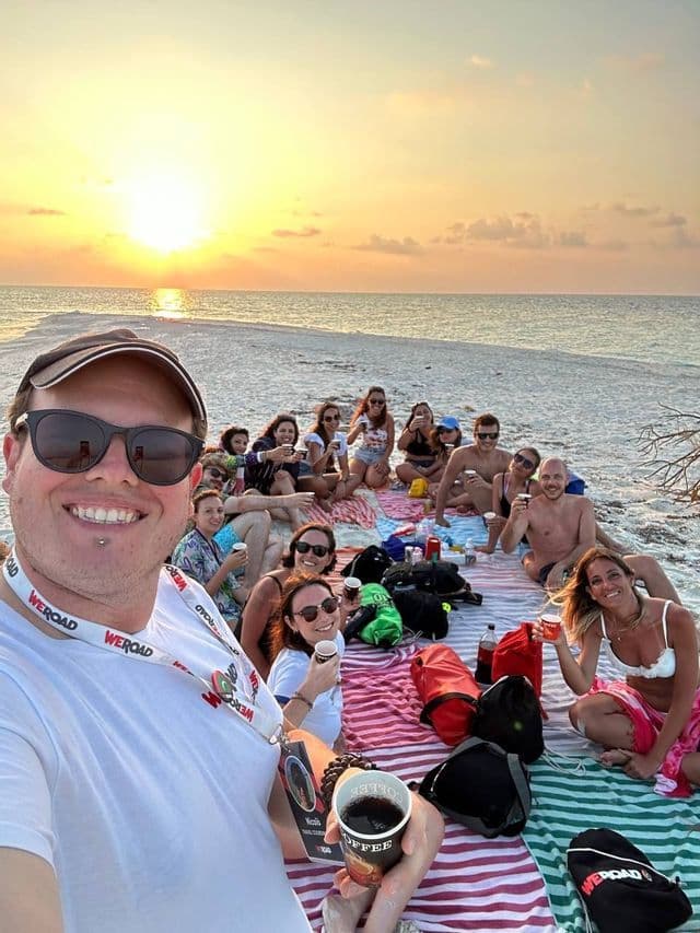 Eine WeRoad-Gruppe macht ein Selfie während eines Picknicks an einem Sandstrand bei Sonnenuntergang.