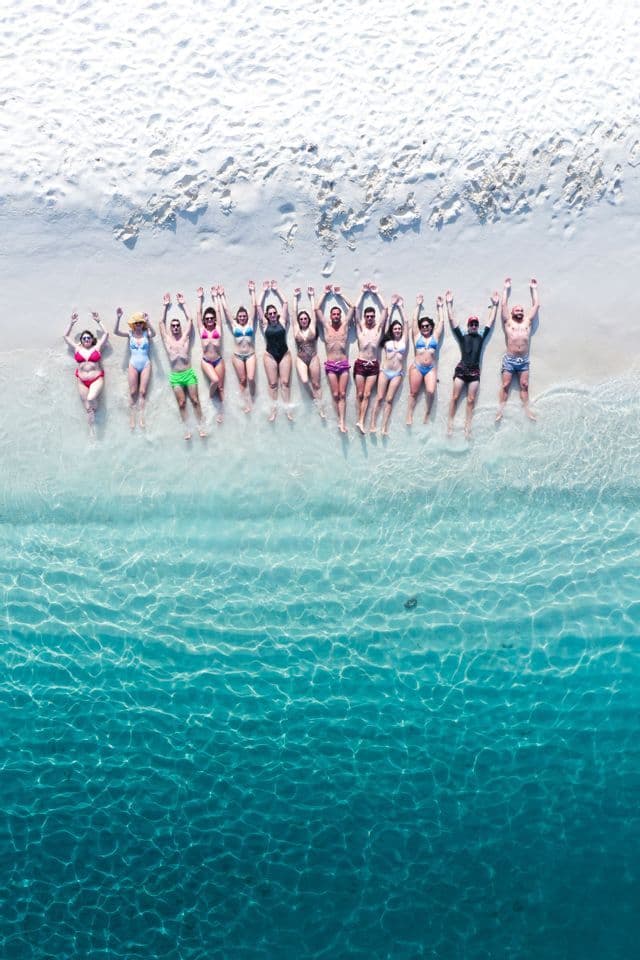 An aerial view of a WeRoad group trip lying in a row on a white sand beach with their arms raised, feet in the clear turquoise water.