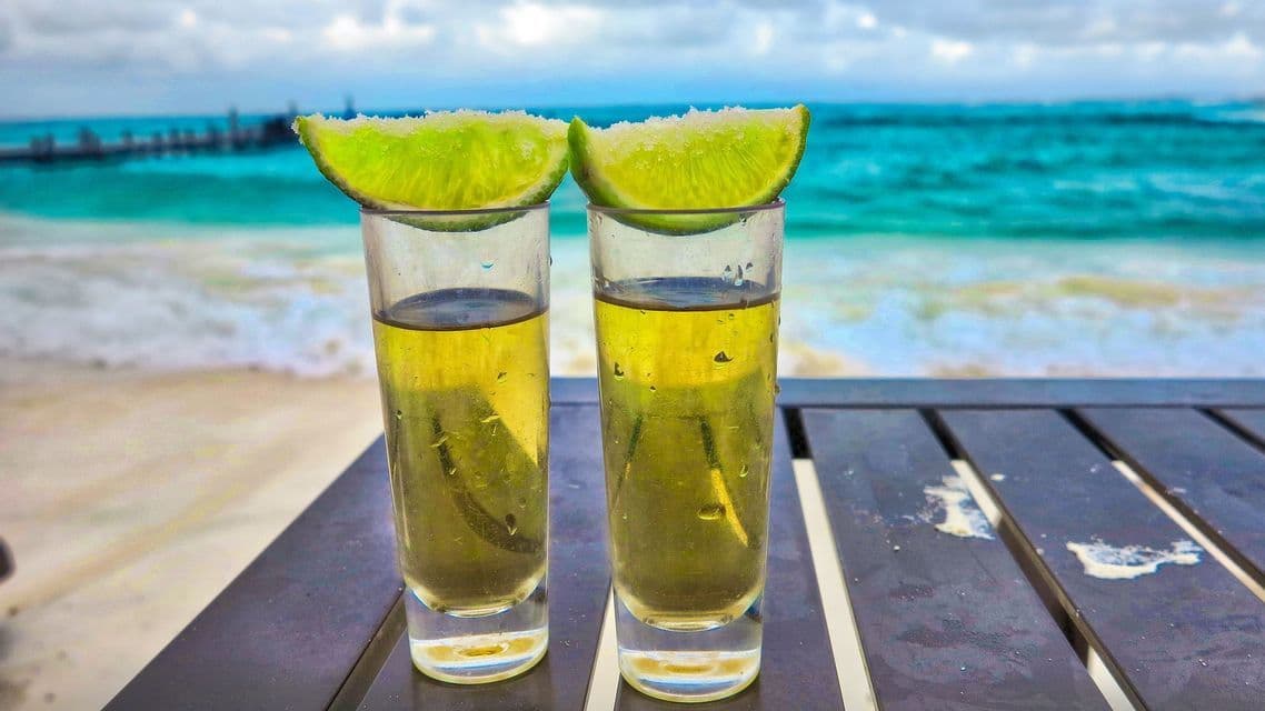 Due bicchierini di tequila con spicchi di lime salati riposano su un tavolo che si affaccia su una spiaggia tropicale con acqua turchese.