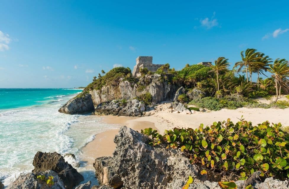 Antiche rovine in pietra si ergono su una scogliera rocciosa sopra una spiaggia sabbiosa con acqua turchese e palme.