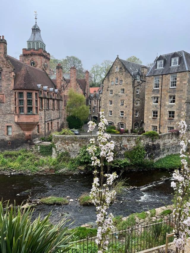 Des bâtiments historiques en pierre, dont un avec une tour horloge, se dressent le long d'une berge verdoyante sous un ciel nuageux.