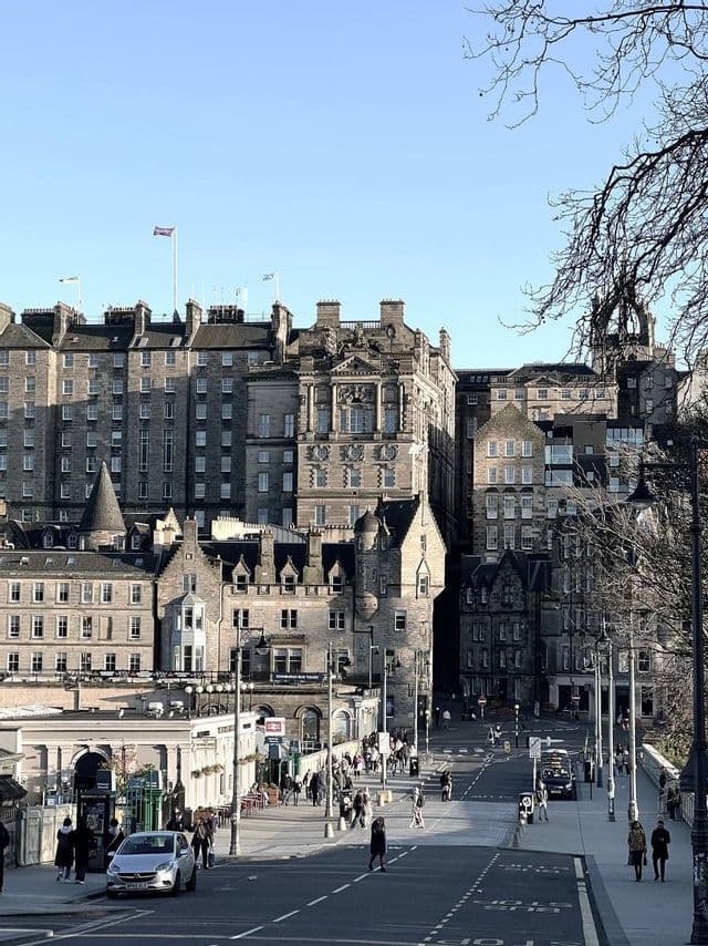 Un'assolata strada cittadina con pedoni che passeggiano lungo storici edifici in pietra sotto un cielo limpido.