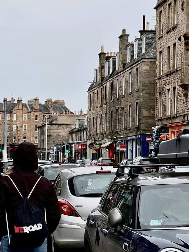 Eine Person mit WeRoad-Rucksack geht eine Stadtstraße entlang, die von Autos und Steingebäuden gesäumt ist.