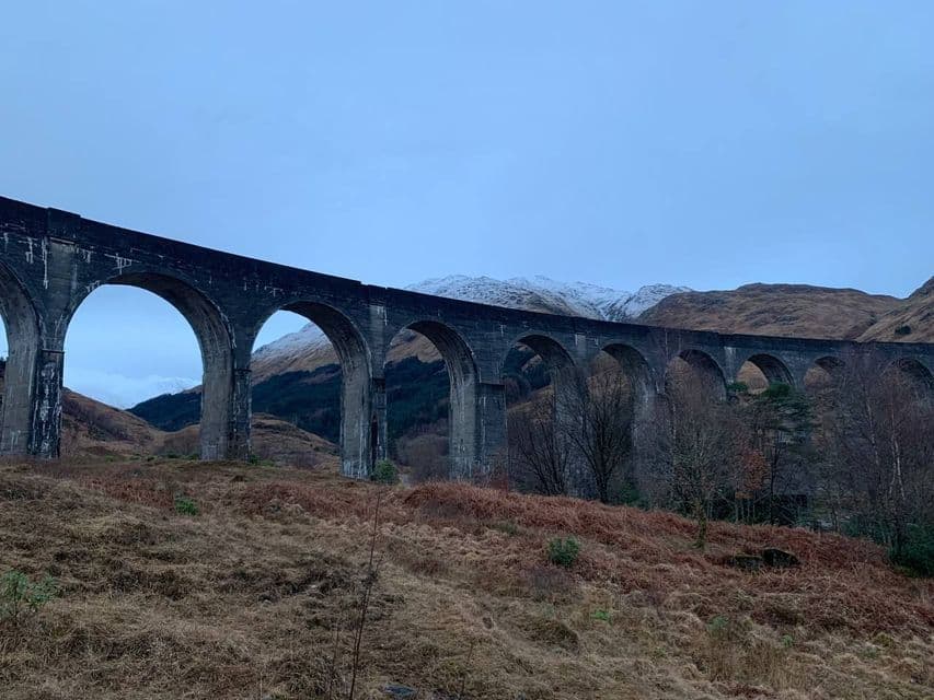 Un lungo viadotto in pietra con più arcate si curva attraverso un paesaggio collinare con montagne innevate sullo sfondo.