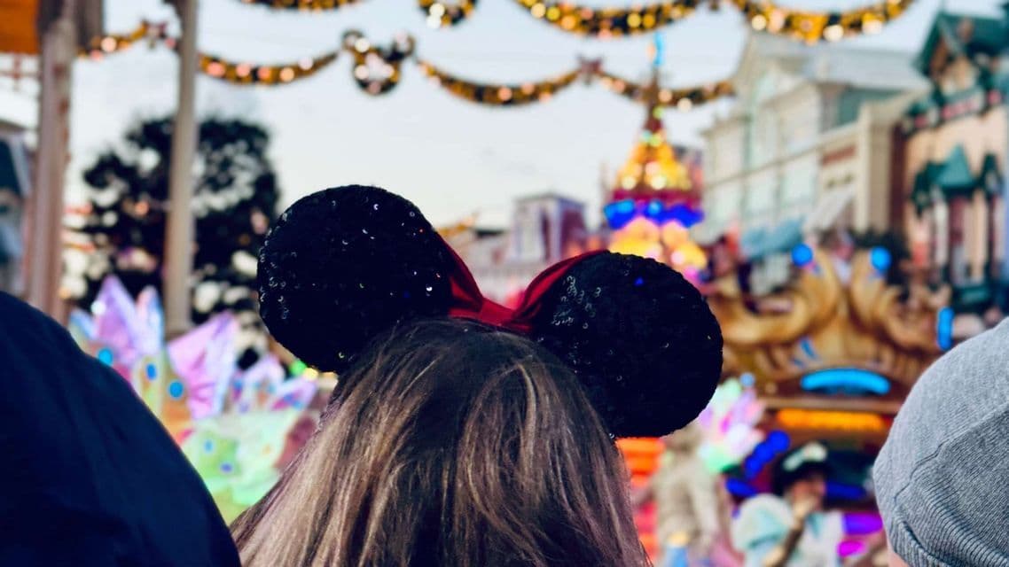 Vista da dietro di una persona con orecchie di Minnie con paillettes nere a una parata colorata con luci di sfondo festive.