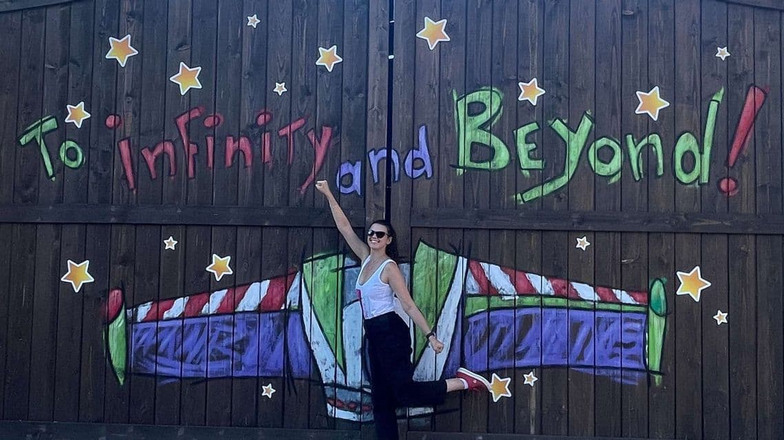 Una donna sorridente posa giocosamente davanti a un muro di legno con graffiti "Verso l'infinito e oltre!".