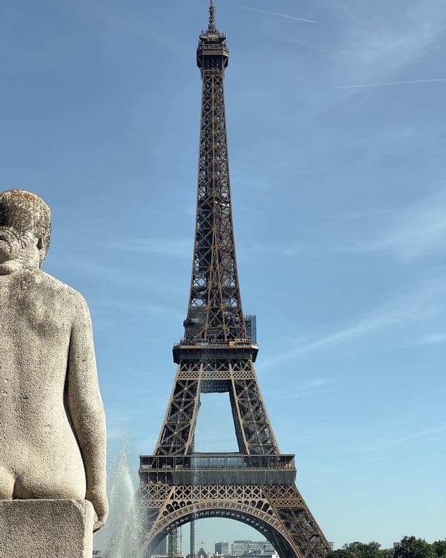 La Torre Eiffel si erge contro un cielo azzurro e limpido, vista da dietro una statua di pietra e una fontana zampillante.