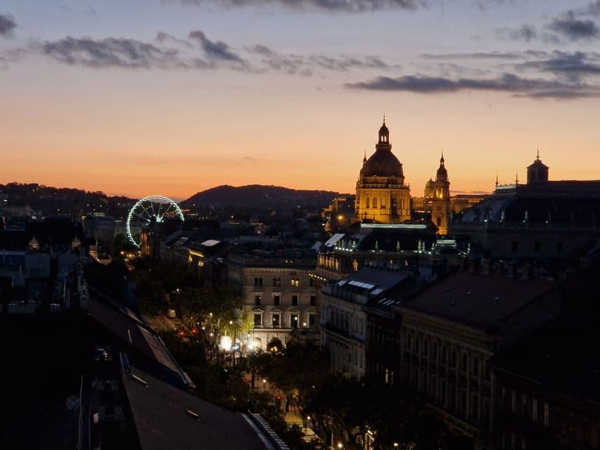 Eine Stadtansicht bei Sonnenuntergang mit einer beleuchteten Basilika und einem großen Riesenrad vor einem orange-lila Himmel.