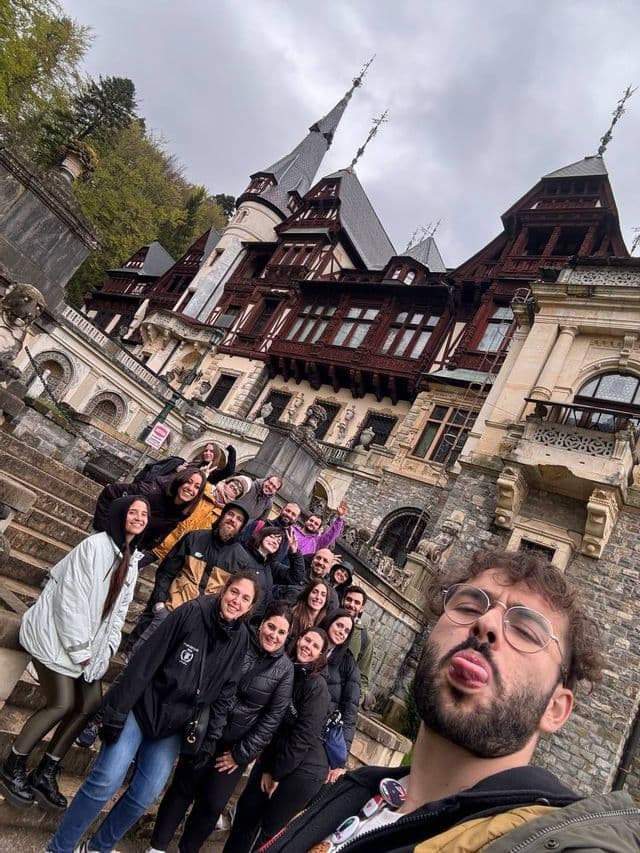 A man takes a selfie with a WeRoad group trip on stone stairs, with a large, ornate castle in the background.