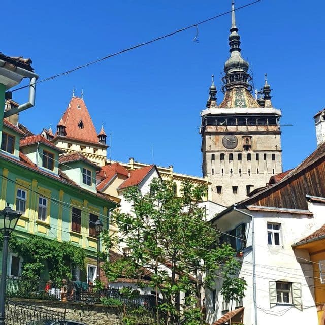 A historic clock tower rises above colorful buildings with red-tiled roofs under a clear blue sky.