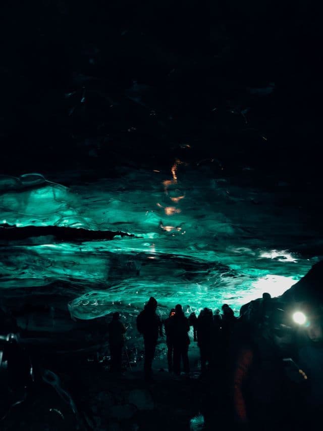 Un viaggio di gruppo WeRoad con persone in silhouette all'interno di una grotta di ghiaccio scura, illuminate da una luce turchese che riflette sulle pareti.
