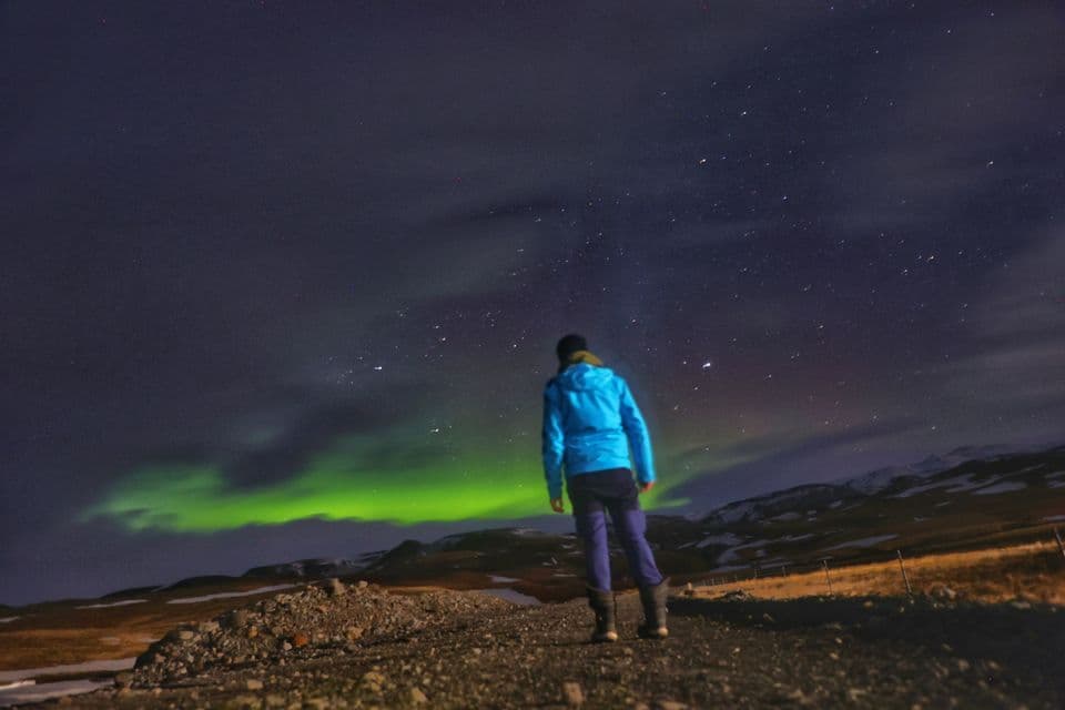 Una persona in giacca blu si trova su un sentiero di notte, osservando l'aurora boreale verde sotto un cielo stellato.