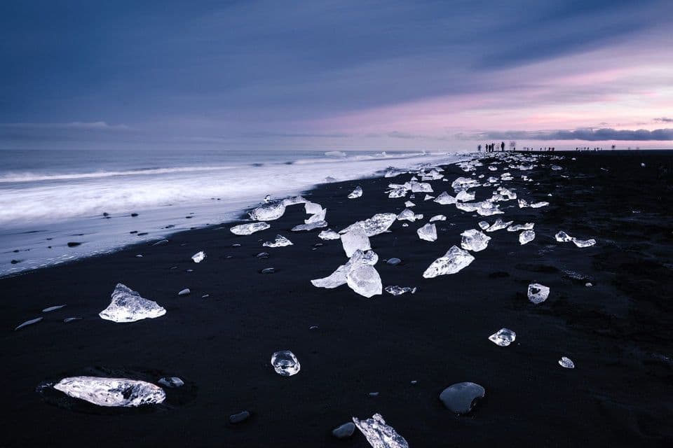 Un viaggio di gruppo WeRoad su una spiaggia di sabbia nera dove blocchi di ghiaccio sono sparsi dalle onde dell'oceano sotto un cielo crepuscolare.
