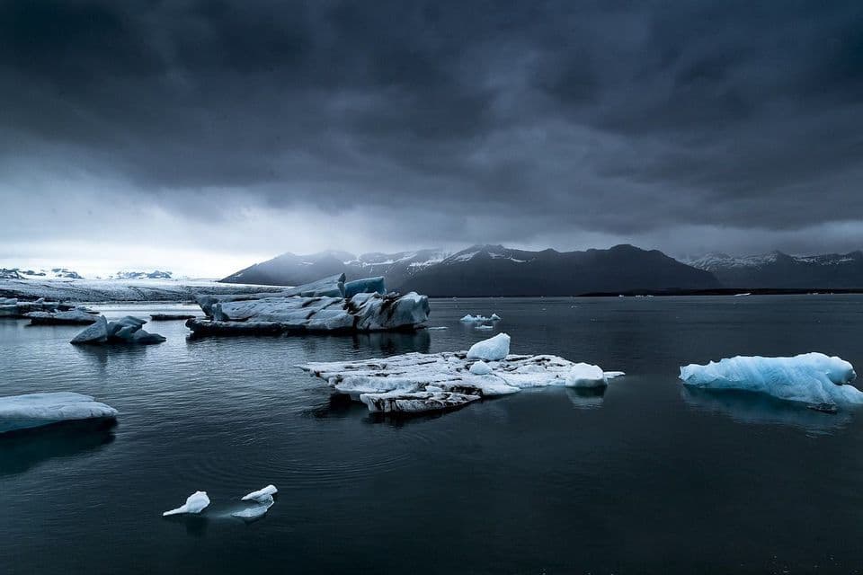 Iceberg galleggiano in una laguna glaciale di fronte a montagne innevate sotto un cielo scuro e nuvoloso, con due uccelli che riposano sul ghiaccio in primo piano.
